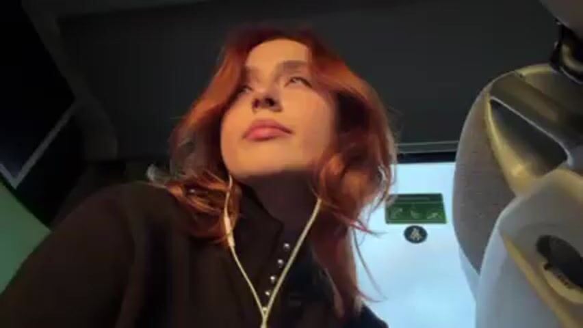 A young woman with red hair is inside a bus or other vehicle. She is wearing a black jacket with silver buttons and white earbuds. She looks around. The bus window is visible with passing scenery and sunlight. There is a sign on the wall in the background. The camera seems to be handheld and slightly unsteady.