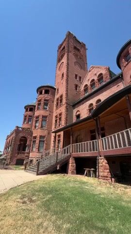 The video shows a large, red brick building with a tower, resembling a castle or an old institution. The building has multiple levels, circular turrets, and arched windows. A wooden staircase leads up to a covered porch with a railing. The building is surrounded by a grassy area, and the sky is clear and blue. The camera pans slowly across the facade of the building, capturing its architectural details.