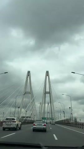 The video captures a drive across a large cable-stayed bridge under a heavily overcast sky. The bridge structure is visible, with its towers, cables, and road surface clearly defined. Cars are seen driving along the multiple lanes in both directions. The perspective is from inside a moving vehicle, likely the driver's point of view. Streetlights line the sides of the bridge, and green road signs indicating directions are visible overhead. The overall tone is neutral, focusing on documenting the experience of crossing the bridge.