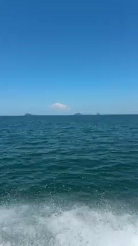 The video shows a view from a moving boat on a sunny day. The sea is dark blue-green and there are white waves breaking near the camera. In the distance, a few islands are visible under a clear blue sky. The boat's frame is visible in the corner.