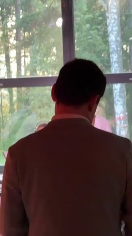 This video shows a band performing. The lead singer is a bald man wearing a white shirt, black tie, and sunglasses. He is playing a red electric guitar and singing into a microphone. Behind him, another man, possibly a bassist, is also playing an instrument. The band is set up in front of a large window that looks out onto a wooded area with trees. The lighting is dim, with red and orange hues visible, suggesting it is nighttime or indoors with stage lighting. In the foreground, the back of someone wearing a suit is visible, indicating they are part of the audience. There are musical instruments, microphones, and music stands present. The overall atmosphere appears to be a live performance at a venue.