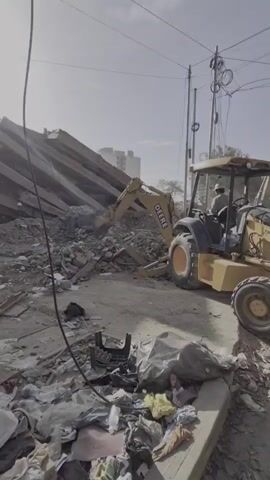 A backhoe loader is being used to demolish a structure. A person wearing a hard hat and work clothes is operating the machinery. The site is littered with debris, including wood, concrete, and trash bags. Power lines are visible in the background, along with buildings. The sky is clear and the sun is shining.