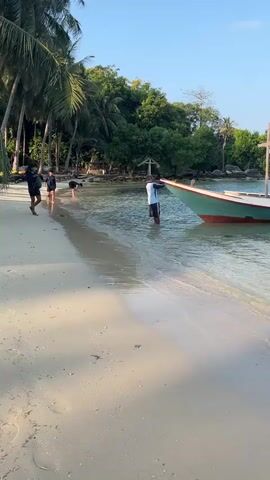The video shows a tropical beach scene with calm, clear water. In the foreground, a sandy beach is visible with a gentle wave lapping at the shore. Palm trees line the left side of the frame, creating a backdrop of lush greenery. In the middle ground, a group of people are interacting near the water's edge. One person stands in the water, appearing to be holding onto a boat. Two other individuals are also in the shallow water. A boat with a red and green hull is partially submerged in the water, its bow pointing towards the shore. The sky above is a clear, light blue. The overall impression is a peaceful, idyllic beach setting.