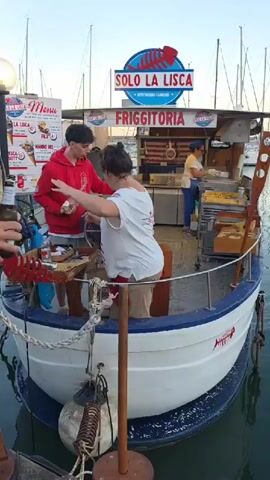 The video shows a food stall on a boat in a harbor. A young man in a red hoodie is handing something to a woman in a white shirt. Another person is working behind the counter, appearing to be cooking. The stall has a sign that says "FRIGGITORIA" and "SOLO LA LISCA". There are boats docked in the background, and buildings along the shore. People are sitting on a dock in the foreground, some eating. The water is calm, and the sky is clear.