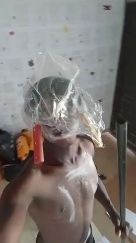 The video shows a person, shirtless and with a clear plastic bag over their head, holding a mop handle. White substance is visible on their face, chest, and the plastic bag. The person moves the mop handle as if brandishing it. The background includes a wall with papers attached, a window, and various household items. The camera is hand-held and the view shaky.