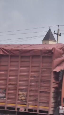 The video begins with a close-up of a large red shipping container with "CAI" in white lettering. The camera then pans to reveal a tall clock tower with a dark pyramidal roof. The clock face shows the time as approximately 12:45. Below the clock, there are decorative arched windows. A sign for "HERITAGE MANSPA" and "SALES OFFICE" with an arrow pointing to the right is visible near the base of the tower. The tower is surrounded by some greenery and streetlights. In the background, industrial buildings and construction cranes can be seen. The sky is overcast.