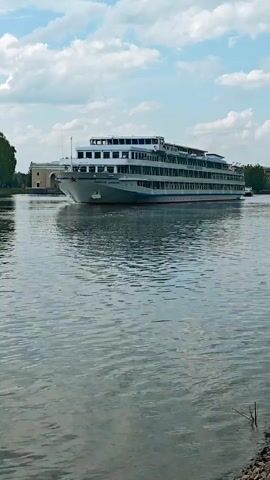 The video depicts a multi-level cruise ship moving on a river. The ship, which appears to be white and gray, has several decks with windows. The river's surface reflects the sky and the ship. In the background, the riverbank is visible, with trees and a building partially hidden by the foliage. As the ship moves out of frame, the camera shifts to the right, revealing more of the riverbank with greenery and some buildings in the distance. The sky is partly cloudy.