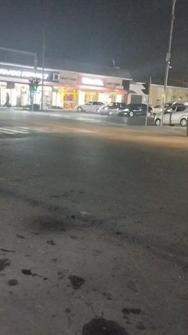 The video captures an urban street scene at night. The pavement in the foreground is dark and uneven, showing some puddles or dark spots.  Across the street, there's a one-story building with businesses, including a fast food establishment called "GAVITA FAST FOOD." There is another business to the left of GAVITA with lit signage. Several cars are parked along the street and one car can be seen driving through the intersection. Pedestrians can be seen crossing the crosswalk in front of the business on the left. Traffic lights are visible at the intersection. The sky is overcast and the lighting is generally dim, illuminated by streetlights and the lights from the businesses.