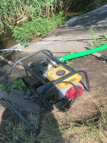 This video shows a portable gasoline-powered water pump operating next to a concrete ditch. The pump is connected to an intake hose that is submerged in the water in the ditch, and an output hose that is draped over the side of the ditch. The pump is powered by a small gasoline engine. The output hose is green and appears to be made of flexible material. The intake hose is brown and ribbed. There is vegetation growing along the edge of the ditch, and dry grass and dirt surround the pump. The scene appears to be outdoors in daylight.