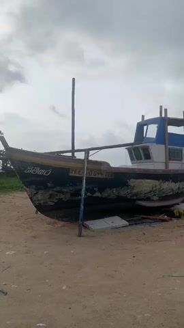 A boat is sitting on a sandy beach under a cloudy sky. The boat is black and white with blue accents and appears to be in disrepair, with peeling paint on its hull. There is a small cabin with windows on the boat. The boat is propped up on supports. Scattered debris, including wood and some yellow fabric, is visible on the sand around the boat. The scene appears to be a coastal area.