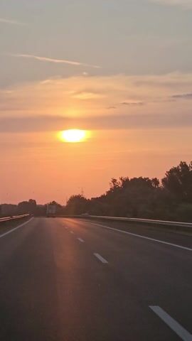 This is a vertical video of a sunset viewed from a moving vehicle on a highway. The sun is setting on the left side of the frame, casting a warm orange glow across the sky and the road ahead. Trees line the right side of the highway, silhouetted against the bright sky. The road itself is asphalt with white dashed lines marking the lanes. The vehicle appears to be traveling at a moderate speed.
