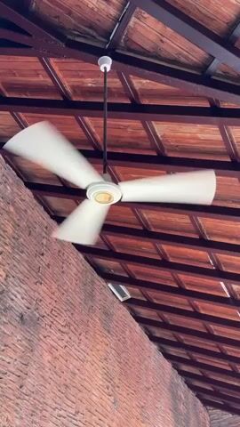 A ceiling fan spins in front of a brick wall and a wooden ceiling. The fan blades are white and appear to be blurred from motion. The ceiling fan is hanging from a rod attached to the ceiling. The ceiling is made of wooden planks and has a grid pattern. The wall is made of red bricks and has some graffiti on it, including a cartoon tiger and a smiley face. The scene is lit by natural light.
