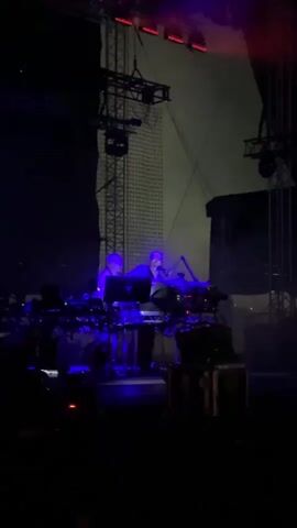 A live musical performance is captured. Two individuals are on a stage, operating electronic music equipment. The stage is dimly lit with vibrant blue and red lights flashing, creating a dramatic atmosphere. Smoke or fog machines are in use, adding to the visual effect. Several pieces of equipment, including synthesizers and mixers, are set up on tables. The background features stage rigging and lighting structures. The perspective is from the audience, looking towards the performers.