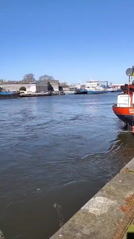 The video shows a waterway with a small boat passing by a docked barge. The barge is blue and has a red crane on it. On the left side of the waterway are buildings and docked boats. The water is murky and reflects the blue sky. On the right is a docked orange and black boat and a stone wall. The sky is clear and blue.