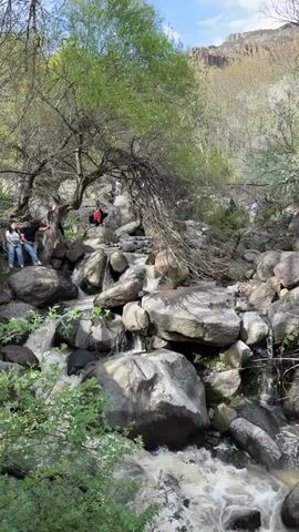 The video shows a rocky river bed with water flowing over large boulders. Several people are visible, some sitting or standing on the rocks near the river, and others walking on the path. The area is surrounded by trees, and a hill or mountain is visible in the background. The sky is partly cloudy.