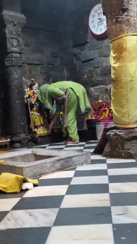 The video shows the interior of a temple. In the foreground, there is a black and white checkered floor. To the left, there is a rectangular stone structure with a yellow cloth draped over its edge. Further back, a woman in a green headscarf and a green and pink floral salwar kameez is standing. She appears to be adjusting her scarf and looking at her phone. Behind her, there is a shrine with idols. To the right of the woman, a man in a white shirt and grey pants is bending over. Further to the right, there is a large yellow cloth hanging and a round clock on the wall showing approximately 1:05. Another person walks into the frame from the left, wearing a purple and blue patterned garment and red pants, with a beige handbag slung over their shoulder. The walls of the temple appear to be made of rough stone.