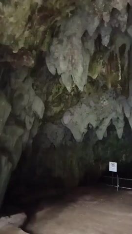 The video shows the interior of a cave. The ceiling is covered in stalactites, some of which are quite large and appear to be damp. The floor of the cave is made of dirt and appears to be a pathway. There are some metal railings and a sign with a white circle and a black line through it, indicating a prohibition. The lighting is dim, with some light sources illuminating the cave walls and stalactites. The overall impression is of a natural cavern, possibly a tourist attraction.