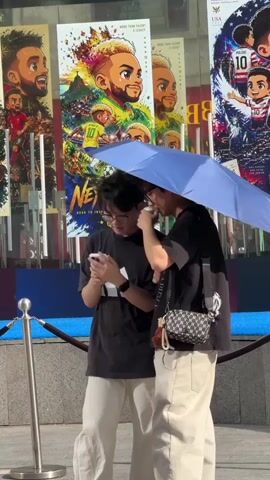 Two young adults, one holding a smartphone and the other an umbrella, stand in front of a building. The person on the left, wearing a black t-shirt with the Adidas logo and beige pants, is looking at the phone. The person on the right, also in a black t-shirt and beige pants, is holding a blue umbrella over both of them, shielding them from the sun, and is looking towards the left. They are standing on a paved surface with a metal stanchion and rope barrier in the foreground.

Behind them, a building facade displays several large posters. The posters feature cartoon-style illustrations of soccer players with vibrant colors and anime-like features. One prominent poster shows a character with blonde hair and a yellow jersey with the number 10. Another poster depicts a group of young boys in soccer uniforms, one with the number 10 on his jersey, with text that includes "USA SOCCER." The text "MORE THAN TALENT" and "A LEGACY" is visible on some posters. The posters suggest a theme related to soccer or sports.

The lighting indicates it is daytime, likely bright and sunny, given the use of the umbrella. The overall scene appears to be outdoors in an urban environment, possibly a commercial or entertainment district. The individuals seem engaged in a casual activity, perhaps taking a selfie or viewing content on the phone. The mood is relaxed and everyday. The location is identified as Xiangzhou, China.