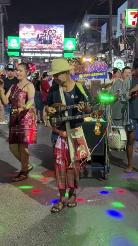 At night, on a pedestrian street in Thailand, a man plays a double-necked guitar while other people dance around him. The man wears a woven hat, a dark shirt, a colorful sarong, and sandals. A young boy in a red and white soccer jersey stands near the camera. In the background is a large electronic billboard that reads "Bangla Walking Street" and several lit store signs. The road is illuminated by multicolored lights. The man's guitar music is amplified through a speaker on a wheeled cart decorated with hanging garlands.