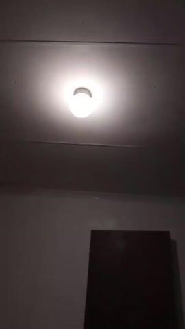 The video shows a room, with a bright light fixture on the ceiling illuminating the space. Below, the wall is visible with a dark, rectangular object, possibly a door or panel, positioned against it. The room is dimly lit except for the area directly around the ceiling light.