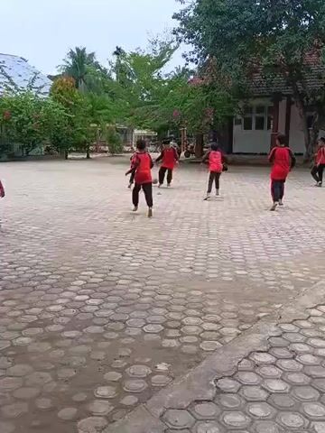 A group of children in red shirts and black pants are playing soccer in a paved schoolyard. The yard is surrounded by trees and buildings. The children are kicking a soccer ball and running around. The weather appears to be overcast.