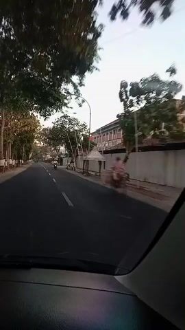 The video is a first-person perspective from inside a vehicle, looking out the windshield. The vehicle is moving along a paved road, with a lane marker visible in the foreground. Trees line the left side of the road, and a motorcycle is visible in the distance on the left. On the right side of the road, there is a long, low building with a white wall and several windows. There are also trees and utility poles along the right side of the road. The sky is overcast. The overall impression is a view of a semi-urban or suburban street scene from a moving car.