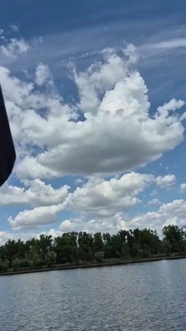 A first-person perspective video shows a boat moving across a body of water, creating a wake. The sky is blue with scattered white clouds. On the left side of the frame, a tree-lined shore is visible, and on the right, the boat's outboard motor is visible. The boat is moving away from the shore and towards the open water.