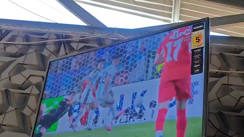 The video shows a close-up of a television screen displaying a soccer match. The screen is filled with two players in red jerseys, one with the number 17 clearly visible on his back. Another player in a white and blue jersey with the number 22 is also visible. The background shows a blurred stadium with spectators. On the right side of the television screen, there is a logo that appears to be for a broadcasting channel, displaying the text "TVRI" and "LANGSUNG" along with a rating of "5". There are also other logos and technical specifications for the TV visible. The image is slightly pixelated and blurry, suggesting it is a recording or a low-resolution stream. The overall environment appears to be indoors, with a patterned wall covering visible behind the television.