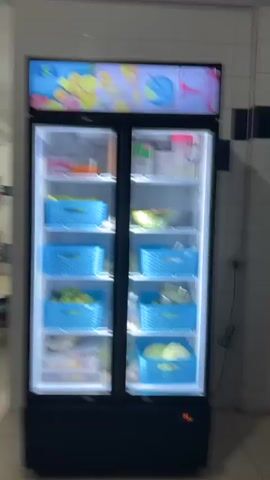 The video shows a professional kitchen. It opens with a shot of a large, double-door refrigerator stocked with produce in blue baskets. The refrigerator is labeled “RSA” at the top with images of fruit. The camera then pans to the left, revealing a large, stainless-steel kitchen. The walls are lined with white tiles accented by a horizontal row of navy-blue tiles. There is a large stainless-steel gas range with a ventilation hood above it. Several stainless-steel tables are arranged throughout the space, some holding kitchen equipment and supplies. A stack of colorful plates is visible on one of the tables. The kitchen appears clean and well-maintained.