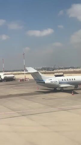 A white private jet with blue accents is parked on an airport tarmac under a partly cloudy sky. The jet is labeled with the registration number 4X-CZH. In the background, other aircraft and airport service vehicles are visible, along with a distant city skyline.