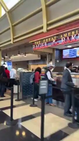 The video shows the interior of what appears to be an airport food court or large cafeteria area. People are standing in lines at various food service counters. Some individuals are wearing casual clothes, while others are in business attire. The environment includes tiled flooring with a black and white checkered pattern and an overhead structure with gold-colored beams. A sign for a food establishment with a red awning is visible.