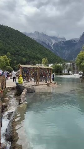 The video opens with a view of a serene, turquoise body of water, possibly a lake or a large pond. In the foreground, a small, man-made waterfall cascades into the main body of water. To the left, a stone pathway lined with wooden railings guides visitors along the water's edge. Further along this path, several people are visible, some walking and others appearing to be taking a break.

Dominating the middle ground is a structure composed of two wooden frames, each densely adorned with numerous colorful padlocks. This display of padlocks suggests a tradition of couples or individuals leaving them as symbols of commitment or wishes. Two individuals are near this structure, one standing to the right and another seated on a large rock to the left.

Beyond the padlock display, a lush green landscape unfolds. A prominent waterfall is seen cascading down a rocky incline, feeding into the water. The backdrop is a majestic mountain range, its peaks partially shrouded by clouds. The mountains are covered in dense green vegetation on their lower slopes, transitioning to rocky, barren summits. Several trees, both large and small, are scattered around the area, adding to the natural beauty. A few more people are visible in the distance, further along the shore of the water, appearing to be enjoying the scenery. The overall atmosphere is one of natural beauty and tranquility, with human elements integrated into the landscape. The sky is overcast, suggesting a cloudy or possibly rainy day.