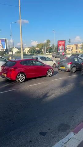 The video shows a street scene with several cars. In the foreground, a red car is visible on the left, and a dark gray car is in the center. A white SUV is parked on the right side of the frame. Behind the cars, a large billboard displays an advertisement with people's faces. A traffic light is visible in the distance. The scene transitions to a closer view of a building with a large Israeli flag flying above it. A white SUV is parked in front of the building, and a person is standing near the car. The building appears to be a commercial establishment with murals painted on the exterior walls. The sky is blue with some clouds.