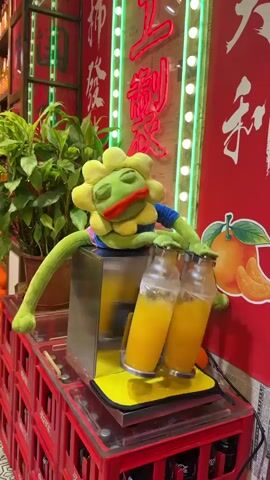 A puppet resembling a frog with yellow flower-like petals on its head is shown operating a juicing machine. The puppet is wearing a blue shirt and is positioned behind the machine, with its arms extended to operate the levers on two bottles filled with orange juice. The juicing machine is metallic with yellow accents. The setup is on top of red plastic crates. In the background, there are green plants and red signage with neon green Chinese characters, along with an image of oranges. The overall environment appears to be a food establishment or market stall. The video captures the action of the puppet seemingly making or dispensing juice.