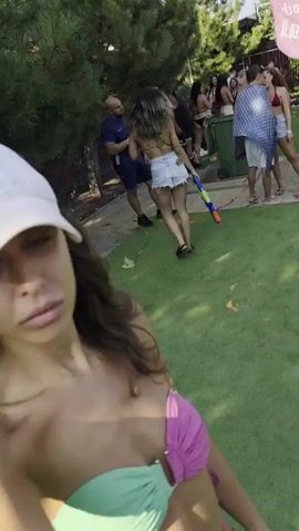 A woman in a white baseball cap and a pink and teal bikini is taking a selfie outdoors. She is smiling and her hand is raised. In the background, there are other people and a green grassy area. Some people are holding what appear to be water guns. There are also scattered colorful objects on the ground. The lighting suggests it is daytime and sunny, with shadows cast on the grass.