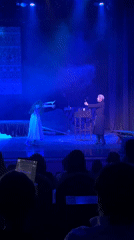 Stage performance features woman in white, magician in blue lighting
