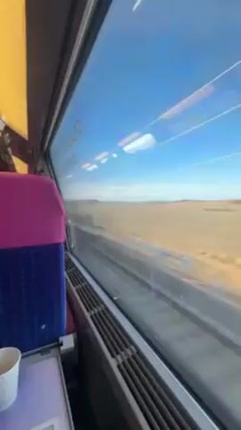 The video shows the view from inside a train, looking out a window at a landscape of dry fields and sparse vegetation under a clear blue sky with some clouds. The train is moving, as indicated by the blurred scenery. The interior of the train car is visible, including a purple and blue seat, a small table, and a heater vent beneath the window. Overhead power lines are visible through the window.