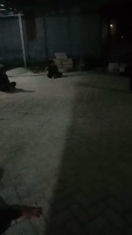 A blurry, low-light video shows an outdoor scene at night. The camera appears to be held by someone walking, as evidenced by the slight shakiness and the ground moving in the foreground. In the background, several figures, likely people, are visible. One figure, dressed in dark clothing, is seen walking away from the camera towards a structure on the left. Another figure, also in dark clothing, appears to be crouched or sitting down near a pile of what looks like bricks or building materials. The environment seems to be a paved area, possibly an alleyway or courtyard, with a wall on the right side. There is some lighting present, casting shadows, but the overall scene is dimly lit. The audio accompanying the video is muffled and unclear, but appears to include speech.