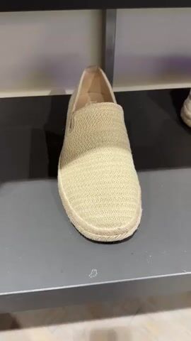 A tan woven TOMS slip-on shoe is seen sitting on a black shelf. A hand picks up the shoe to show the label and the side.