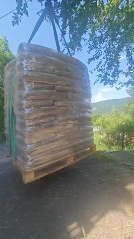 A pallet of bagged materials, shrink-wrapped and secured with green strapping, is being lifted by a crane. The bags appear to be light brown or tan in color with darker speckles. The pallet is made of wood. The scene is outdoors with bright sunlight filtering through the leaves of a tree in the foreground. In the background, there are hills and some greenery. The sky is blue. The video appears to be a close-up shot of the lifting operation.