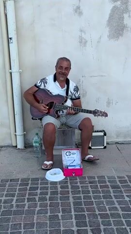 A man is playing an acoustic guitar while sitting on a stool. He is wearing a white polo shirt with a black and white pattern on it and grey shorts. He has a grey beard and is smiling. In front of him, on the cobblestone ground, is a white hat, an open pink box with a sign that reads "CD 10€ 29 Titres" and a bottle of water. To his right is a small amplifier. The man is sitting against a light-colored, textured wall, with a white pipe running vertically on the left side of the frame. The scene appears to be outdoors, possibly on a street or promenade, and the lighting suggests it is daytime. The man's posture and smile convey a sense of enjoyment and engagement with his music or surroundings.