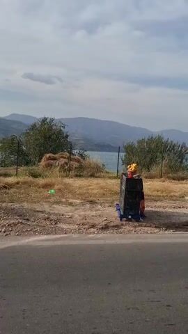 A video captures a view of a body of water, likely a lake or reservoir, with mountains in the background under a cloudy sky. In the foreground, a road is visible, bordered by a concrete barrier. The area between the road and the water appears to be dry, with some sparse vegetation and a fence. Trees are scattered throughout the scene. The camera is moving, likely from a vehicle, as evidenced by the slight motion blur and the changing perspective. No people are visible in the video.