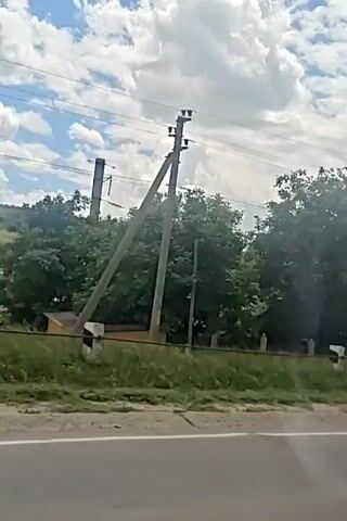The video shows a view from a vehicle moving along a road. Power lines and poles are visible alongside the road, with trees and vegetation in the background. The sky is cloudy with patches of blue. Several houses and buildings can be seen nestled among the trees. The sun is shining, and the weather appears to be warm. The video pans slightly as the vehicle continues its journey.
