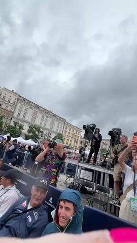 This is a video of an outdoor event in a city square, possibly Krakow, Poland given the text "Kraków" visible on some of the barriers. The sky is overcast, suggesting a cloudy or potentially rainy day, with several people using umbrellas and wearing hooded jackets. The scene is crowded, with an audience seated in rows of chairs, facing a stage area where media personnel with cameras and video equipment are set up on platforms. Some individuals in the audience are holding up their phones, likely taking pictures or videos of the event. Photographers with professional cameras are also present, actively capturing images. The background features historic architecture, typical of European cities, with grand buildings and tents that might indicate vendor stalls or event infrastructure. The atmosphere appears to be one of anticipation for an event.