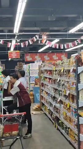 The video shows the interior of a brightly lit supermarket aisle. Three individuals, appearing to be store employees based on their maroon and white uniforms, are gathered around a shopping cart. One employee is standing on a small stepladder, reaching up to a shelf, while another employee is leaning over the shopping cart, seemingly assisting or observing. The third employee stands nearby, also looking towards the shelf. The shelves are stocked with various packaged goods, including snacks and other food items. Red and white flags are strung across the ceiling, suggesting a festive or national occasion. The overall environment suggests normal store operations.