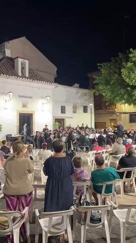 A nighttime outdoor concert takes place in a plaza. A large orchestra, comprised of men and women in dark attire, is seated on chairs and stands. A conductor in a dark suit directs the orchestra. An audience of people, many of whom are seated in white plastic chairs, watches the performance. Some audience members are standing. Buildings with lit windows and decorative lighting surround the plaza. A large tree with green foliage is visible in the background. The sky is dark.