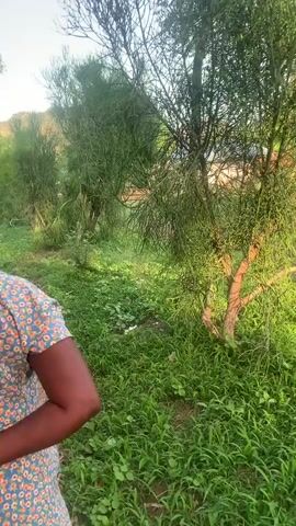 A person wearing a floral print dress walks away from the camera down a dirt path. The person is holding a tan envelope behind their back. The path is surrounded by green grass and trees. The video is taken during the daytime.
