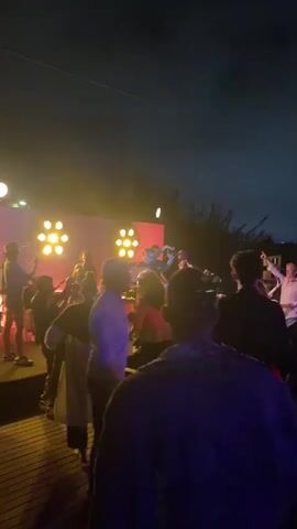 The video shows a group of people gathered outdoors at night. They appear to be at a party or event, with music playing and people dancing. There are colorful lights illuminating the scene, casting a pink and yellow glow on the crowd. Some people are holding drinks, and the overall atmosphere seems lively and festive. The sky is dark, indicating it is nighttime.