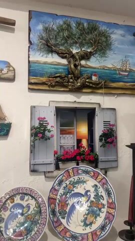 The video shows a wall display of decorative items, possibly in a shop. The wall is white and textured. On the wall are several framed or mounted artworks and decorative objects. In the upper portion of the frame, there's a painting of an olive tree with a textured trunk, set against a seascape with a sailboat. To the left of this, there's a smaller wooden artwork depicting a sailboat. Below the olive tree painting is a faux window with blue shutters, revealing a sunset scene. In front of the window are potted plants with pink flowers. On a shelf below the window, there are two large decorative plates with colorful bird and floral designs. To the left of these plates, on a lower shelf, are various ceramic items including small jars with lids, a pitcher, and a small dish. Further to the left, a tall, decorative candlestick with a floral pattern is visible. To the far left of the frame, a blue decorative piece resembling a door or window frame with a painted scene of a sailboat and a moonlit sky is mounted on the wall. There is also a vertical wooden artwork depicting a mermaid. The lighting appears to be artificial, casting some shadows. The items suggest a theme of maritime and Mediterranean decor.