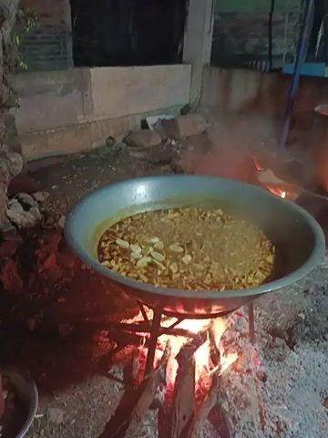 A high-angle shot reveals a person adding food from a small bowl into a large metal pot that is suspended over a wood fire. The fire is blazing, emitting smoke, and is surrounded by pieces of wood. The person is wearing blue pants and sandals, and only their legs and feet are visible. The food being added to the pot appears to be chunks of something, possibly vegetables. The pot contains a liquid with floating pieces. To the left of the scene is a concrete structure, and the ground is covered with dirt and small rocks. The camera angle is tilted slightly downward, focusing on the cooking process.
